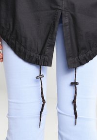 Haut en tissu noir avec un ourlet arrondi, doté d'un cordon de serrage réglable à la taille. Associé à un jean skinny bleu clair.