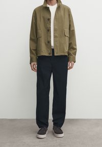 Olivgrön bomullsjacka med krage, knäppning, två framfickor, kombinerad med marinblå vida byxor och grå skor.