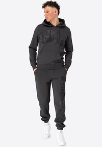 Ensemble en sweat à capuche et pantalon de jogging gris foncé, avec une impression de logo sur la poitrine et sur la jambe. Le tissu semble lisse et texturé. Les accents incluent une poche kangourou et des poignets élastiques.