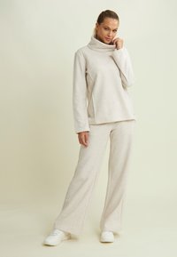 Top in fleece beige, morbido, con ampio collo a scaldacollo e cuciture angolate, abbinato a pantaloni larghi coordinati e sneakers bianche.