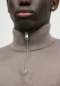 Taupe värvi kampsun kõrge kraega, millel on keskelt hõbedane lukku. Sile tekstuur ja soonikuga krae detail.