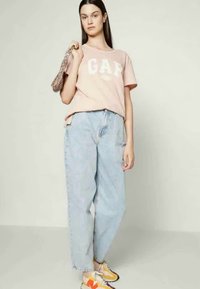 T-shirt rosa claro com o logótipo "GAP", jeans de corte relaxado e sapatilhas multicoloridas. Modelo transporta uma carteira de padrão.