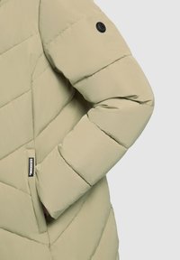 Chaqueta acolchada color oliva claro con paneles cosidos, un botón de logo negro en la manga y un bolsillo lateral con una etiqueta de marca.