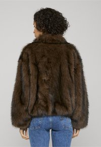 Brown faux fur jacket with a high collar and wide sleeves, featuring dark stripes, worn over blue denim jeans. Textured, soft appearance.