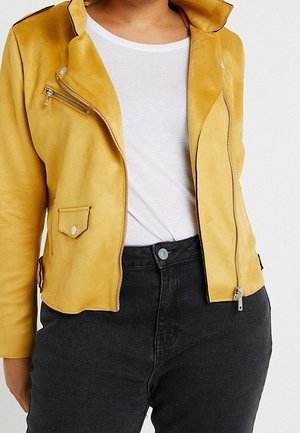 Frau trägt eine senfgelbe Jacke mit Reißverschluss über einem weißen Hemd und hoch taillierte schwarze Hose, steht vor einem schlichten Hintergrund.