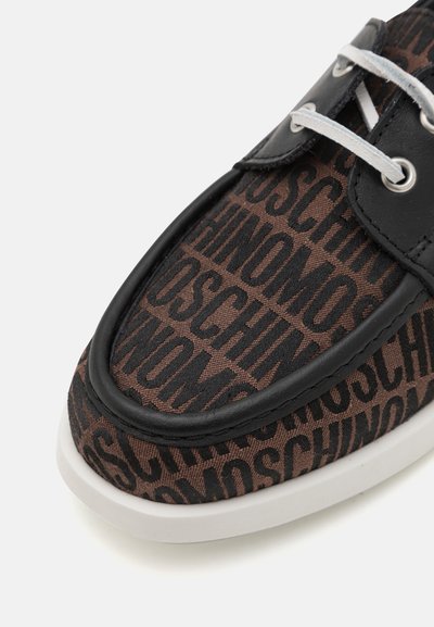 Zapatilla de tela marrón con un destacado patrón de logo negro, contrastada por acentos de cuero negro y suela de goma blanca.