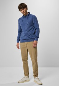 Modrá mikina s dlouhým rukávem a kapucí vyrobená z jemné látky s vysokým límcem, kombinovaná s béžovými chinos se zrolovanými manžetami a bílými teniskami.