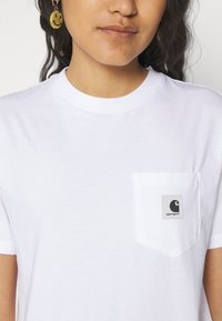 Hvit bomull t-skjorte med rund hals og korte ermer. Har en lomme på venstre bryst med en grå lapp som viser Carhartt-logoen.