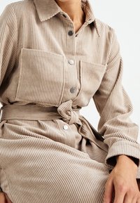 Abito camicia in corduroy rigato beige con colletto, bottoni frontali, due tasche sul petto e una cintura legata in vita per un aspetto aderente.