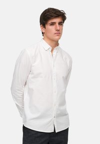 Chemise boutonnée blanche en tissu léger, avec des manches longues, un col classique et un petit logo sur la poitrine.