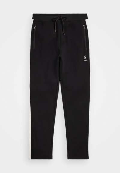 Svarte joggebukser laget av mykt materiale, med snøring i livet, glidelåslommer og en hvit logo på venstre underbein.