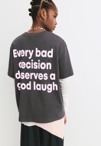 Persoon met gevlochten haar draagt een donkergrijze shirt met grote roze tekst op de achterkant die leest: "Elke slechte beslissing verdient een goede lach."