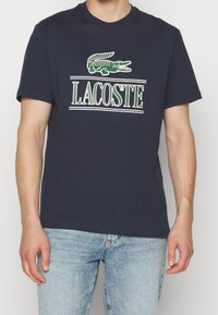 Man wearing a navy blue Lacoste t-shirt with a green crocodile logo and white text, paired with light blue jeans.