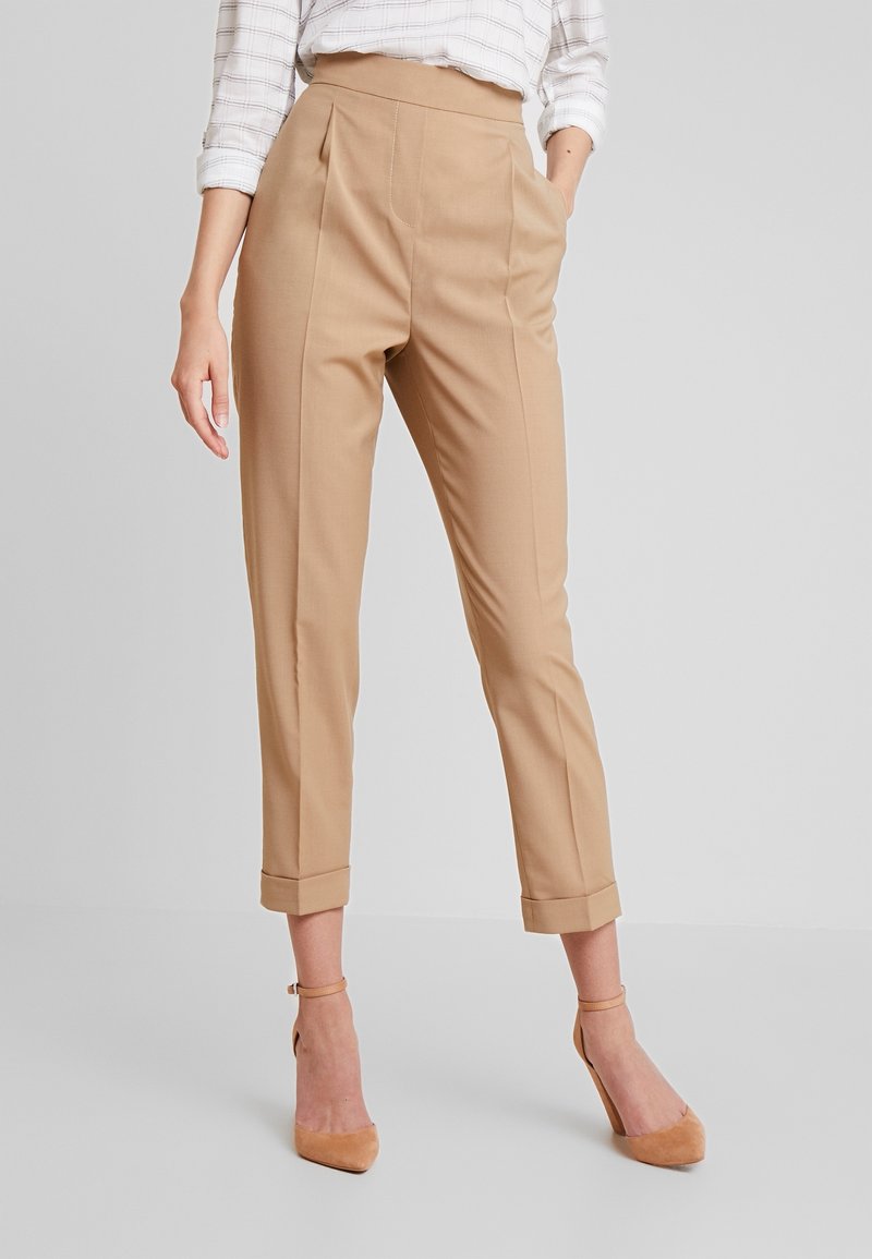 Femme portant un pantalon beige slim fit raccourci avec des revers, associé à des talons beige à bride cheville et une chemise blanche à carreaux aux manches retroussées.