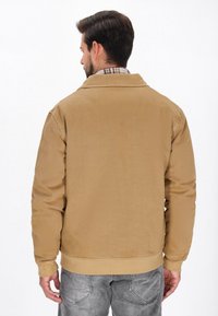 Veste en velours côtelé beige avec un design à col, présentant une finition texturée et un ourlet à côtes, portée sur une chemise à carreaux et un jean gris.