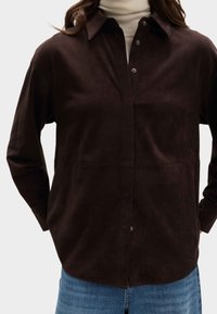 Hnědá semišová košile s límcem, knoflíkovým zapínáním a dlouhými rukávy. Má strukturovaný povrch a volnější střih, doplněná modrými denimovými džíny.