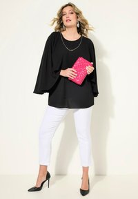 Haut noir à manches longues avec des manches fluides, pantalon blanc court, et escarpins noirs. Tenant une pochette rose matelassée. Fond uni.