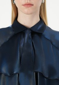Blouse en satin bleu marine avec un col classique, dotée d'une fermeture à nouer au cou et de détails volantés sur le devant, texture lisse.