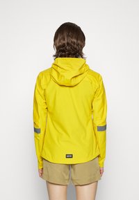 Keltainen vedenpitävä takki hupulla, jossa heijastavia yksityiskohtia hihoissa ja logo helmassa. Sileä pinta, istuva malli.