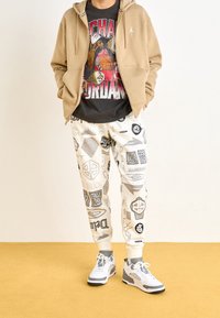 Beżowa bluza rozpinana na zamek, czarny t-shirt z grafiką, białe spodnie dresowe z czarnymi i szarymi logo oraz białe trampki z szarymi akcentami.