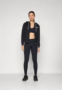 Zwarte hoodie met ritssluiting en trekkoorden, gecombineerd met een zwarte cropped top en leggings met hoge taille. Model draagt zwarte sportschoenen.