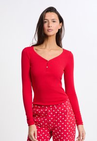 Červený dlouhý rukáv top s výstřihem do V, zdobený detaily knoflíků. Měkká, pružná žebrovaná tkanina. Spárováno s červenými kalhotami s vzorem.