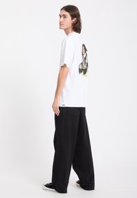 Jeune homme aux cheveux châtain moyen porte un t-shirt blanc à motif graphique, un pantalon large noir et des baskets noires, debout de profil sur fond blanc.
