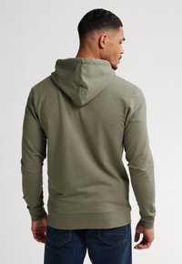 Felpa con cappuccio di colore verde oliva, con maniche lunghe, vestibilità ampia e polsini a costine. Il tessuto appare morbido e con una texture liscia.