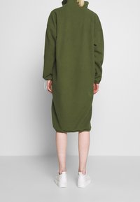 Överdimensionerad, grön fleeceklänning med hög krage, långa ärmar och elastiska ärmmuddar. Bärs med vita sneakers som når till mitt på vaden.