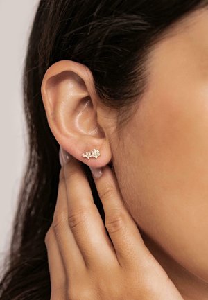 Goldene Ohrstecker mit kleinen, klaren Steinen, die in einem floralen Muster angeordnet sind. Nahaufnahme eines Ohrs und einer Hand, die sanft das Ohrläppchen berührt.