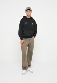 Haut à capuche noir avec poche avant, texte du logo vert ; pantalons beiges texturés ; baskets blanches avec accents gris ; personne se tient contre un fond blanc.