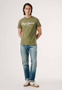 Olivengrønn t-skjorte med hvit tekst "Pepe Jeans London", kombinert med lyseblå jeans og hvite joggesko. Enkel design, korte ermer.