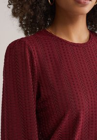 Bordo teksturirana majica s uzorkom cik-cak. Ima okrugli izrez i blago napuhane duge rukave, izrađena od mekog materijala.