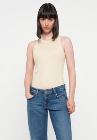 Beige ribbet tank topp med høy hals og uten ermer, kombinert med blå høyhalsede jeans med lommer og knappeåpning.