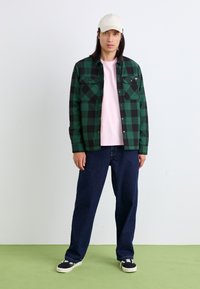 Chemise à carreaux verte et noire avec deux poches poitrine, superposée à un t-shirt rose clair. Associée à un jean bleu foncé et des baskets noires.