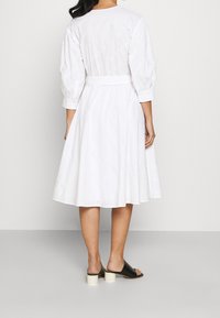 Robe blanche jusqu'aux genoux avec des manches bouffantes, taille cintrée et détails en dentelle. Associée à des chaussures à enfiler noires avec un talon bloc.