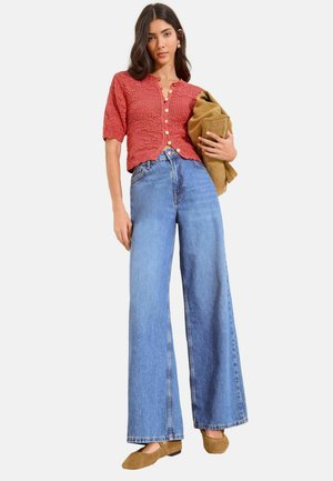 Frau trägt korallenrotes, kurzes Strick-Oberteil mit Knöpfen, hoch taillierte, weit geschnittene blaue Jeans, braune Ballerinas, hält eine beige Jacke vor weißem Hintergrund.