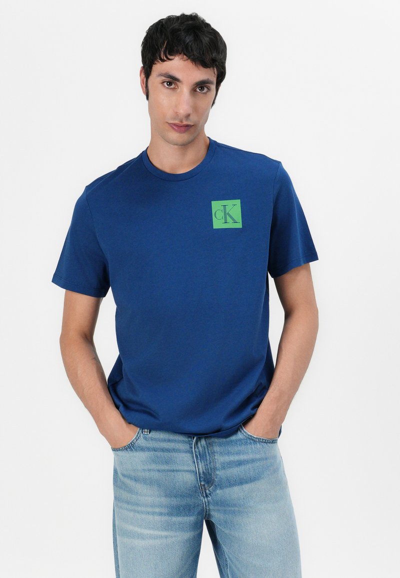 Granatowa bawełniana koszulka z krótkim rękawem i okrągłym dekoltem z zielonym kwadratowym logo po lewej stronie klatki piersiowej, zestawiona z jasnoniebieskimi dżinsami.