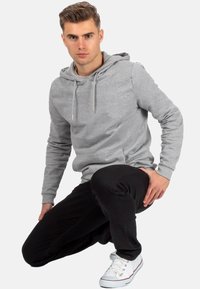 Haut en gris avec cordon de serrage, poche avant ; associé à un pantalon de survêtement noir et des baskets blanches. Modèle posé en position accroupie.