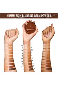 Yummy Skin Blurring Balm Powder i en rund, bronsefarget beholder. Prøver på armer viser 11 nyanser, som varierer fra lys til mørk, med en jevn tekstur.