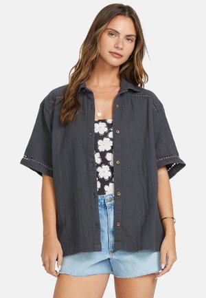Chemise boutonnée gris foncé à manches courtes, avec une bordure en dentelle sur les épaules et une coupe décontractée, associée à un haut floral noir et à des shorts en denim.