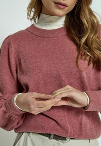 Pink knitted sweater with ribbed neckline and cuff details, featuring a soft texture. Model layered it over a white high-neck top.