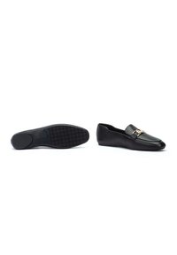 Mocasines de cuero negro con punta redonda, que presentan un detalle de hardware en tono dorado en la parte frontal y una suela de goma texturizada.