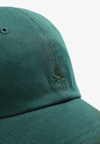 Groene katoenen pet met een gestructureerd oppervlak, voorzien van een geborduurd doodskop en kruisbeenderen logo in een iets donkerder groen.