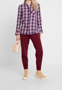 Rutig skjorta i marinblått och rosa, långa ärmar, knäppning fram; matchad med burgundy joggers och beige hög-top sneakers. Bär en rosa handväska.