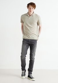 Polo shirt beige a maniche corte con colletto, abbinato a jeans grigi aderenti e scarpe da ginnastica nere con suole e lacci bianchi.