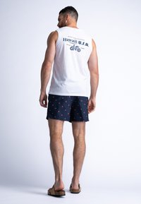Maglietta bianca senza maniche con stampa "Hawaii U.S.A." sul retro, abbinata a pantaloni da bagno blu navy con piccoli motivi floreali rossi.
