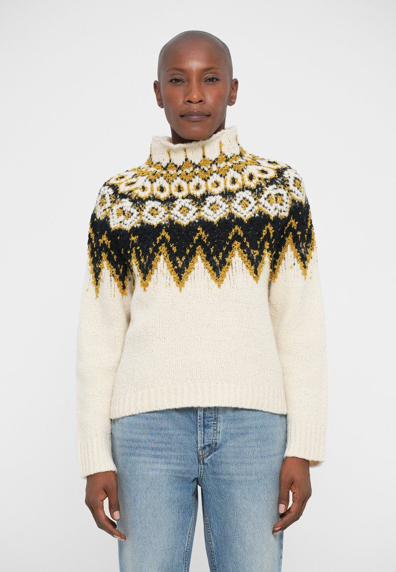 Persona che indossa un maglione di pelliccia color crema con motivi geometrici neri e gialli, abbinato a jeans azzurri chiari su uno sfondo neutro.