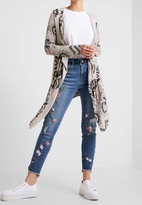 Gebreide lange cardigan met zwart bloemenpatroon, wit T-shirt, versleten blauwe jeans met kleurrijke bloemenborduursels en witte sneakers.