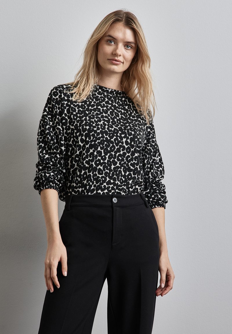 Schwarzer und weißer Leopardenmuster-Sweatshirt mit langen, leicht puffierten Ärmeln. Kombiniert mit hoch taillierten schwarzen Hosen mit Knopfverschluss.